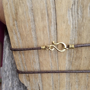 May include: Close-up of a brown cord necklace with a gold-colored hook clasp. The necklace is displayed against a weathered wooden background, highlighting the contrast between the materials and textures.