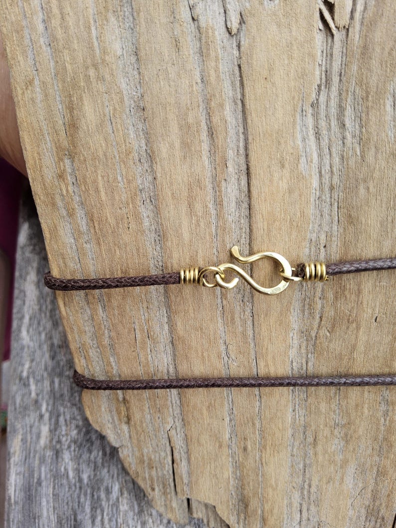 May include: Close-up of a brown cord necklace with a gold-colored hook clasp. The necklace is displayed against a weathered wooden background, highlighting the contrast between the materials and textures.
