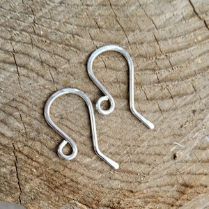 Puede incluir: Dos ganchos de pendientes plateados se muestran sobre una superficie de madera marrón texturizada. Los ganchos tienen un diseño simple y curvado con un pequeño bucle en la parte superior y un extremo recto y puntiagudo. Probablemente son para joyería hecha a mano.