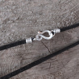 Pu&ograve; includere: Primo piano di una collana con cordino nero e chiusura color argento. La chiusura &egrave; a gancio e occhiello, e il cordino sembra essere fatto di un materiale intrecciato. La collana &egrave; esposta su una superficie di legno invecchiato.