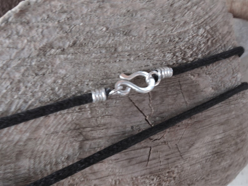 Pu&ograve; includere: Primo piano di una collana con cordino nero e chiusura color argento. La chiusura &egrave; a gancio e occhiello, e il cordino sembra essere fatto di un materiale intrecciato. La collana &egrave; esposta su una superficie di legno invecchiato.