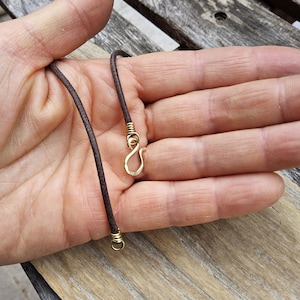 May include: A brown cord necklace with gold-colored metal accents. The necklace features a hook clasp and is displayed against a wooden background.