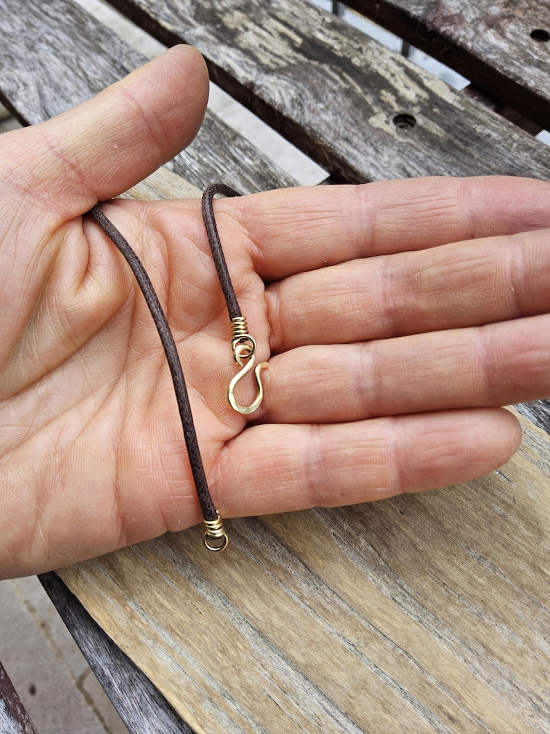 May include: A brown cord necklace with gold-colored metal accents. The necklace features a hook clasp and is displayed against a wooden background.