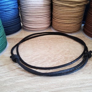 Könnte beinhalten: Ein schwarzes Kordelarmband mit verstellbaren Knoten liegt auf einer hellen Holzoberfläche. Im Hintergrund sind Spulen mit farbigem Kordel in Grün-, Blau-, Beige- und Brauntönen zu sehen, was auf Bastelbedarf hindeutet.