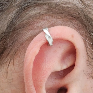 Puede incluir: Un ear cuff de tono plateado con un diseño trenzado similar a una cuerda. La banda de metal tiene un acabado liso y pulido, y se coloca en el cartílago superior de la oreja, creando una apariencia de bucle continuo alrededor del borde.