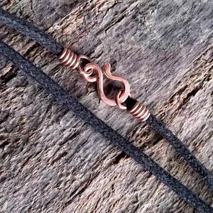 Pu&ograve; includere: Primo piano di una collana con un cordino grigio scuro e una chiusura a gancio color rame. La chiusura ha la forma di una S ed &egrave; avvolta con filo di rame. Lo sfondo &egrave; una superficie di legno strutturata e invecchiata.