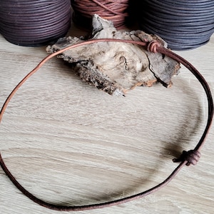 Puede incluir: Un cordón de cuero marrón con un nudo en cada extremo. El cordón está sobre una superficie de madera clara.