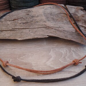 May include: A brown and black leather cord necklace with four knots. The necklace is lying on a piece of driftwood.