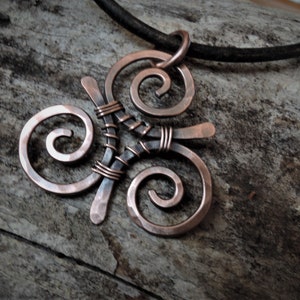 Può includere: Un ciondolo in rame con un design a tripla spirale, che presenta una texture martellata e un accento di filo nero. Il ciondolo è attaccato a un cordino in pelle nera.