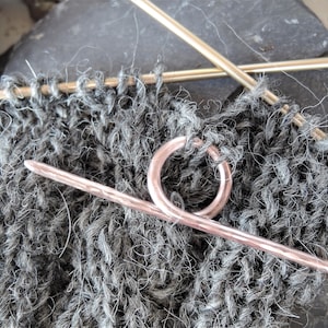 Op de afbeelding: Een close-up van een roosgoud breinaald met een lus aan het einde, gebruikt om steken op hun plaats te houden tijdens het breien. De naald ligt op een stuk grijs gebreid stof.