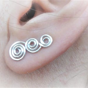 May include: Silver spiral earrings with three connected coils. The earrings are shown in a person's ear.