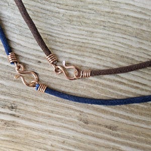 Pu&ograve; includere: Primo piano di due collane con cordino scamosciato e chiusure color rame. Una collana &egrave; blu navy, l'altra &egrave; marrone. Le chiusure hanno la forma di un gancio e di un occhiello.