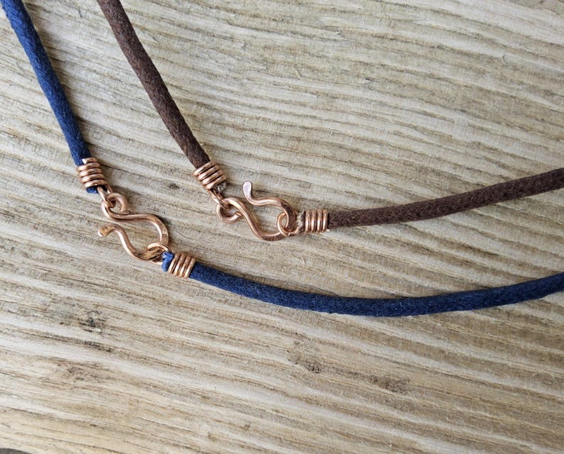 Pu&ograve; includere: Primo piano di due collane con cordino scamosciato e chiusure color rame. Una collana &egrave; blu navy, l'altra &egrave; marrone. Le chiusure hanno la forma di un gancio e di un occhiello.