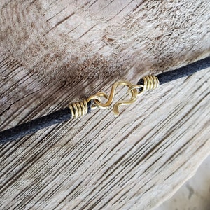Pu&ograve; includere: Primo piano di una collana con cordino nero e chiusura a gancio color oro. La chiusura ha la forma di un gancio a S. Il cordino &egrave; attaccato alla chiusura con un filo color oro. Lo sfondo &egrave; una superficie di legno marrone chiaro.