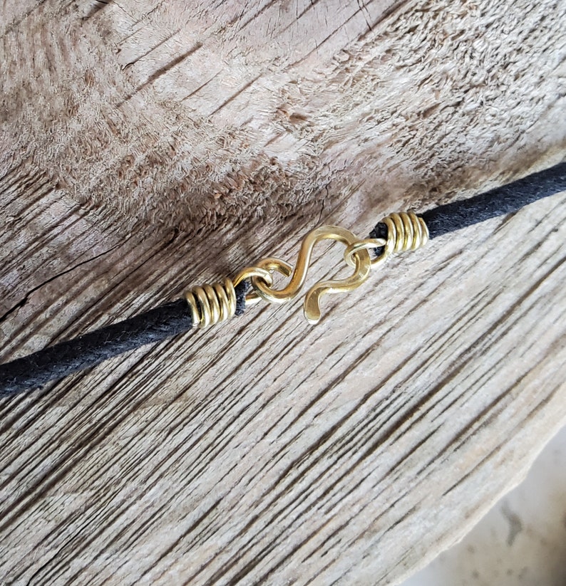 Pu&ograve; includere: Primo piano di una collana con cordino nero e chiusura a gancio color oro. La chiusura ha la forma di un gancio a S. Il cordino &egrave; attaccato alla chiusura con un filo color oro. Lo sfondo &egrave; una superficie di legno marrone chiaro.
