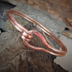 Può includere: Un braccialetto in rame con un design annodato. Il braccialetto è realizzato con due pezzi di filo di rame che sono intrecciati insieme per formare un anello.