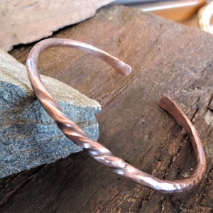 May include: A handcrafted copper cuff bracelet with a twisted design. The open-ended bracelet has a hammered texture and is displayed on a wooden surface. The bracelet's diameter is approximately 6.35 cm.