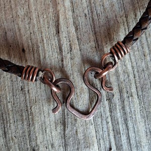 May include: A brown leather cord necklace with a copper clasp. The clasp is a simple, handmade design with a heart shape.