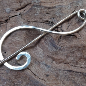 May include: A silver brooch with a hammered finish and a curved design. The brooch has a pin that goes through a loop at the top.