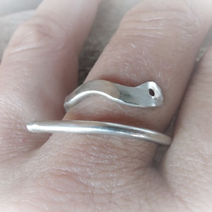 May include: A silver ring with a unique, flowing design. The ring features a smooth, curved band that wraps around the finger, with an abstract, wave-like element on top. The top element has a small hole. The ring is made of a shiny, metallic material.