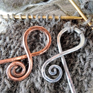 May include: Close-up of two handcrafted shawl pins. One is copper-colored, the other is silver-colored. Both pins have a hammered texture and a decorative swirl design. They rest on a gray knitted fabric, with knitting needles and yarn in the background.