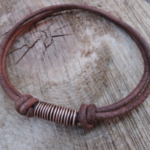 May include: A brown leather bracelet with a copper coil accent. The bracelet is tied with a knot and has a rustic look.