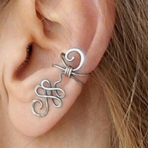 May include: Silver ear cuff with a delicate, intricate design. The cuff is made of wire and features a spiral and a knot.