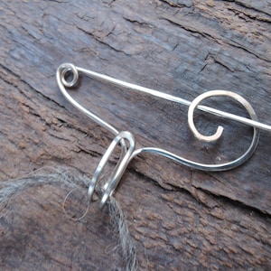 May include: A silver metal brooch with a spiral design. The brooch is attached to a piece of brown yarn.