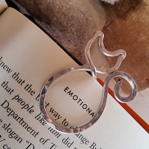 Op de afbeelding: Zilveren metalen bladwijzer in de vorm van een opgerolde kat. De bladwijzer ligt op een boek met het woord "EMOTIONAL" zichtbaar op de pagina.