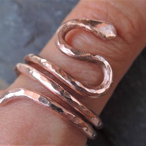 Könnte beinhalten: Ein kupferfarbener Ring in Form einer Schlange, die sich um einen Finger windet. Der Ring hat eine gehämmerten Textur, mit dem Schlangenkopf oben. Der Ring wickelt sich mehrmals um den Finger.