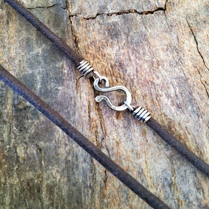 Pu&ograve; includere: Primo piano di una collana con un cordino marrone scuro e una chiusura a gancio color argento. La chiusura ha la forma di una S ed &egrave; attaccata al cordino con filo avvolto. Lo sfondo &egrave; una superficie di legno invecchiato.