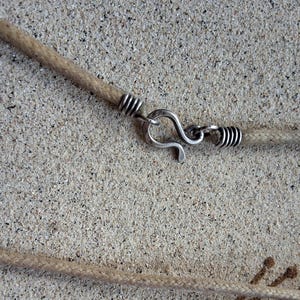 Pu&ograve; includere: Primo piano di una collana fatta a mano con un cordino di corda beige e una chiusura a gancio color argento. La chiusura &egrave; collegata al cordino con un avvolgimento di filo d'argento. La collana &egrave; esposta su una superficie strutturata marrone chiaro.