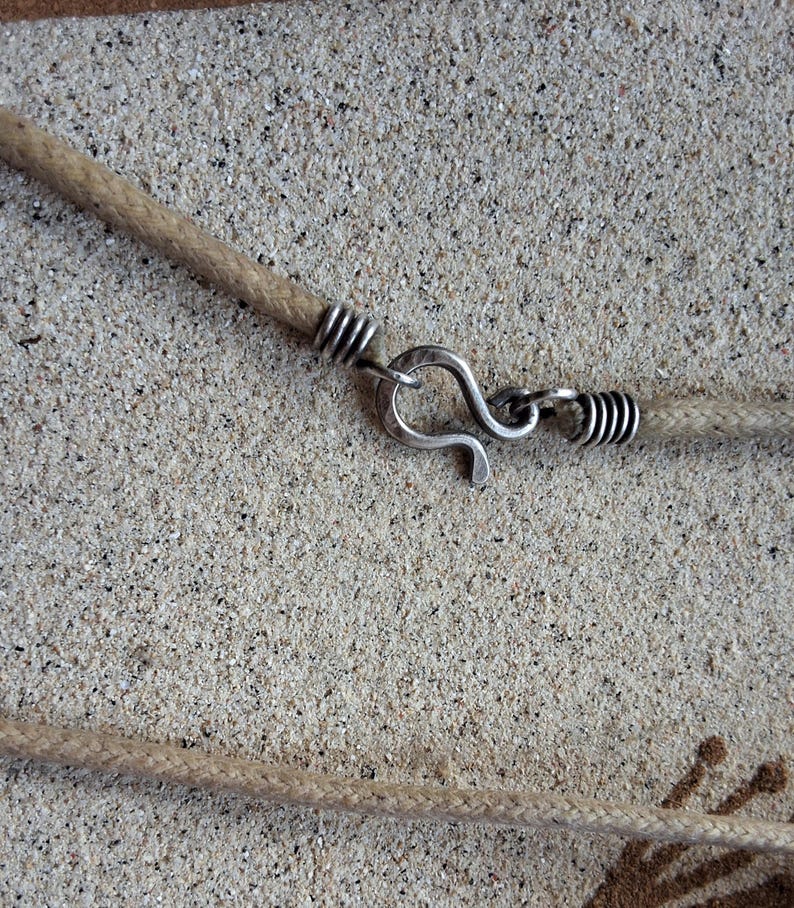 Pu&ograve; includere: Primo piano di una collana fatta a mano con un cordino di corda beige e una chiusura a gancio color argento. La chiusura &egrave; collegata al cordino con un avvolgimento di filo d'argento. La collana &egrave; esposta su una superficie strutturata marrone chiaro.