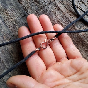 Pu&ograve; includere: Primo piano di una collana con cordino nero e chiusura a gancio color rame. La chiusura &egrave; attaccata al cordino con filo di rame. La collana &egrave; esposta su una superficie di legno strutturata.