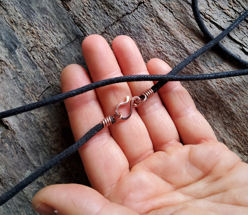 Pu&ograve; includere: Primo piano di una collana con cordino nero e chiusura a gancio color rame. La chiusura &egrave; attaccata al cordino con filo di rame. La collana &egrave; esposta su una superficie di legno strutturata.