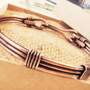 May include: A copper-colored bracelet with a hook clasp. The bracelet is made of three parallel, curved wires, with wrapped wire detailing at the ends and center. The bracelet is displayed in a cardboard box.