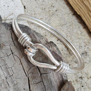 May include: A silver-colored hook and eye bracelet. The bracelet features a hook closure and is made of two parallel bands. The hook and eye closure is wrapped with wire. The bracelet is displayed on a weathered wooden surface.