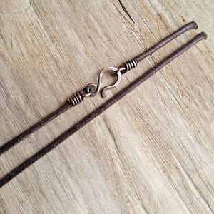 May include: A close-up of a brown necklace with a copper-colored hook clasp. The necklace cord is dark brown and appears to be made of a woven material. The clasp is a simple hook design, and the necklace is set against a light brown wooden surface.