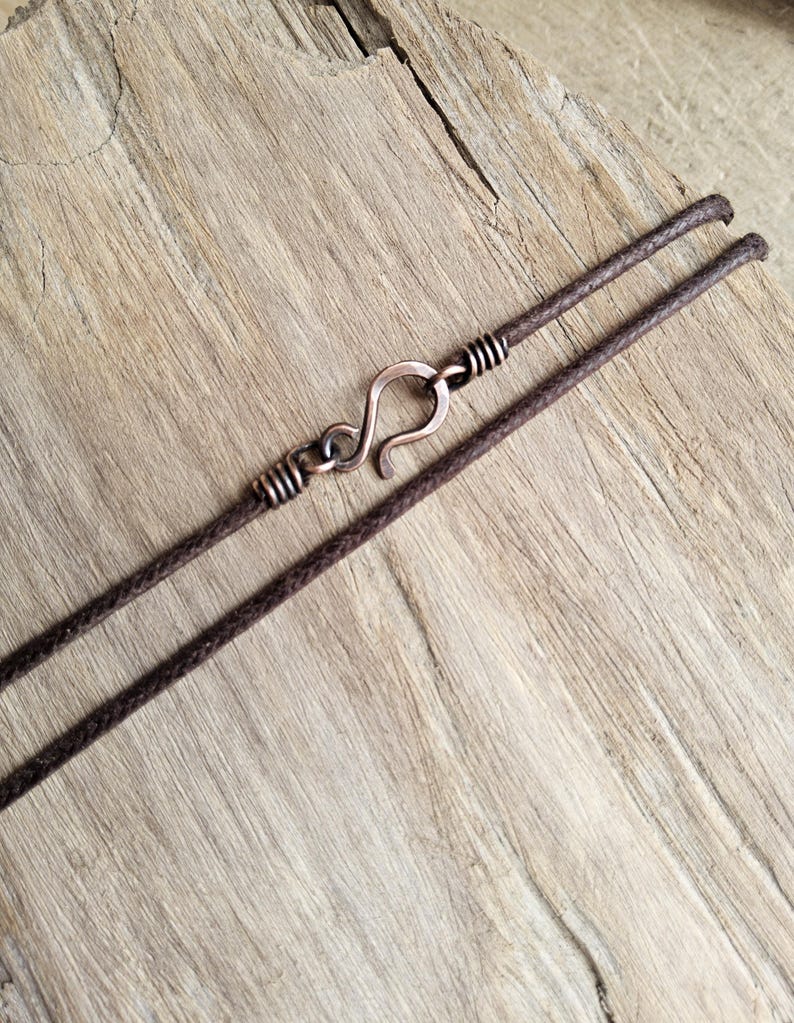 May include: A close-up of a brown necklace with a copper-colored hook clasp. The necklace cord is dark brown and appears to be made of a woven material. The clasp is a simple hook design, and the necklace is set against a light brown wooden surface.