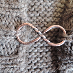 May include: A close-up of a brown knitted fabric with a twisted metal clasp in the shape of an infinity symbol.
