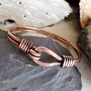 May include: A copper bracelet with a unique clasp design. The bracelet is made of two strands of copper wire that are twisted together to form a loop. The clasp is made of a single strand of copper wire that is bent into a hook shape.
