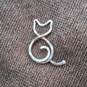 Puede incluir: Una escultura de alambre en forma de gato de plata con textura martillada. La escultura está enroscada en una posición sentada con la cola envuelta alrededor de su cuerpo.