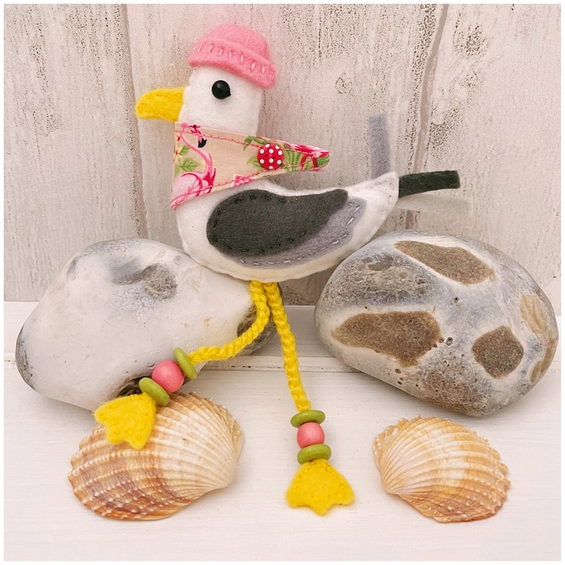 May include: A felt seagull toy wearing a pink hat and a floral scarf. The seagull is perched on a gray rock with two seashells in front of it. The seagull has a yellow cord with pink and green beads hanging from its legs.