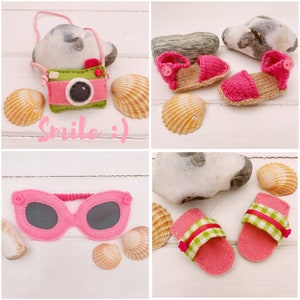 May include: A set of felt crafts featuring a pink camera with the text "Smile :)", pink sunglasses, and pink and green sandals. The crafts are arranged on a white wooden background with seashells.