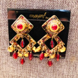 Puede incluir: Un par de pendientes colgantes con un patrón de leopardo en forma de diamante, un estrás rojo central y múltiples cuentas rojas colgantes y pequeños dijes circulares dorados. Los pendientes están sujetos a una tarjeta negra con la etiqueta mamó Designs.