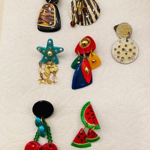 May include: A collection of seven assorted clip-on earrings featuring diverse shapes and motifs, including a turquoise starfish with a seahorse charm, a watermelon slice design, cherry-like red discs, and various geometric patterns in bold colors.