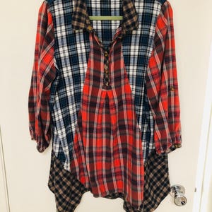 May include: A long-sleeved plaid shirt in a patchwork design. The shirt features a mix of red, blue, white, and brown plaid patterns. It has a collared neckline and button details down the front. The hemline has an asymmetrical cut.