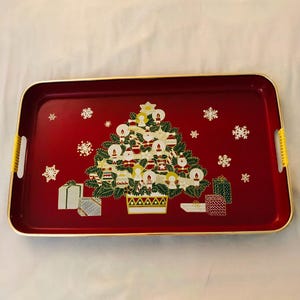 Puede incluir: Una bandeja de servicio rectangular roja con borde dorado y asas laterales amarillas. El centro presenta un gráfico de un árbol de Navidad decorado con adornos, ángeles y regalos, rodeado de patrones de copos de nieve blancos sobre fondo rojo.