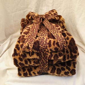 Peut inclure: Une pile de serviettes pliées présentant un motif léopard marron et beige. Les serviettes sont attachées ensemble avec un ruban assorti à motif léopard, noué en un nœud au centre supérieur de la pile, maintenant les couches de tissu pliées.