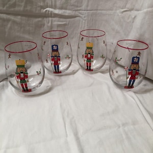 Op de afbeelding: Set van vier heldere, stelenloze wijnglazen met een rode rand. Elk glas heeft een kleurrijke illustratie van een notenkrakersoldaat met een zuurstok. Feestelijk vakantieontwerp met peperkoekmannetjes en hulst.